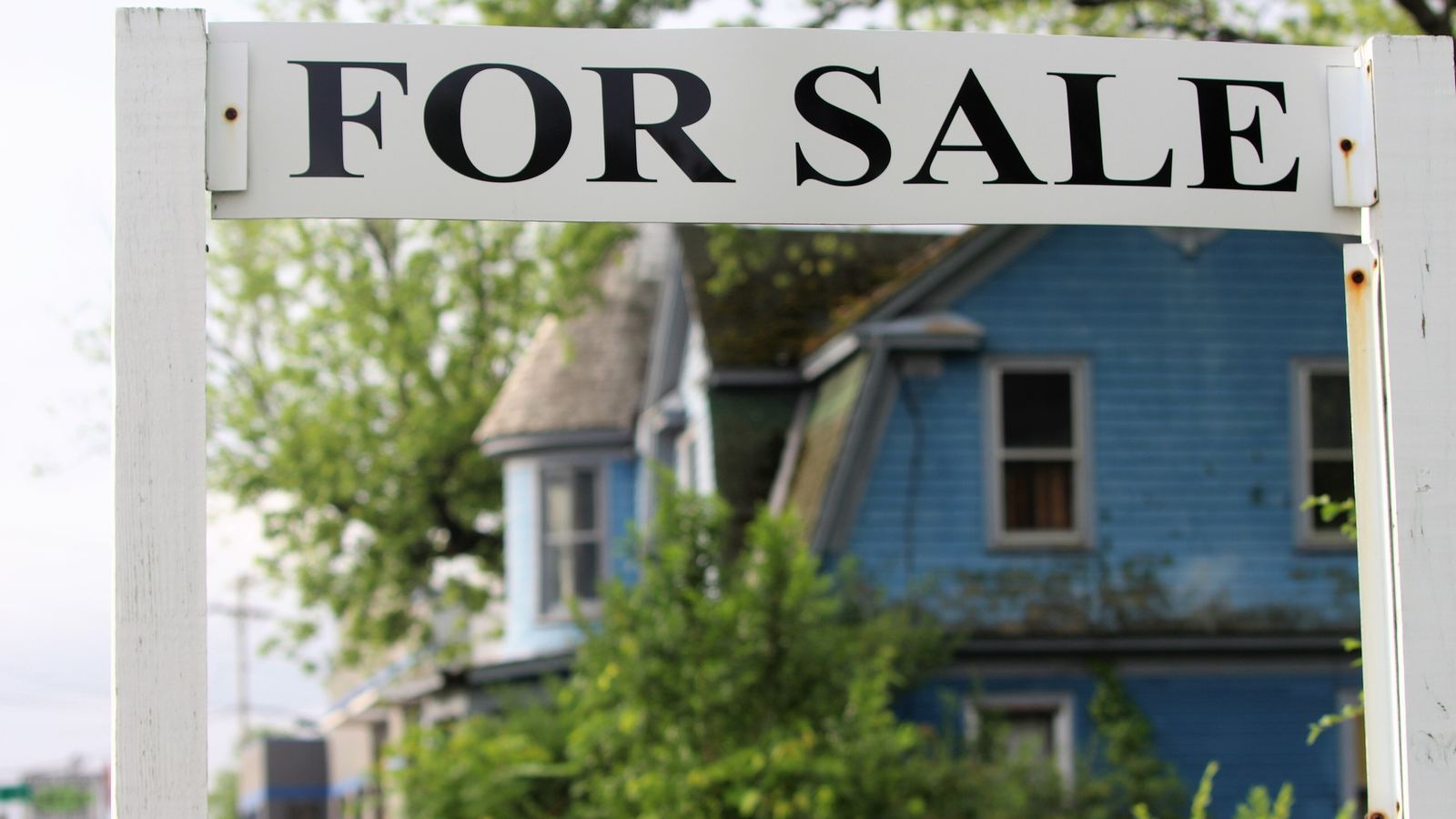A large 'For Sale' sign in front of a vacant blue house