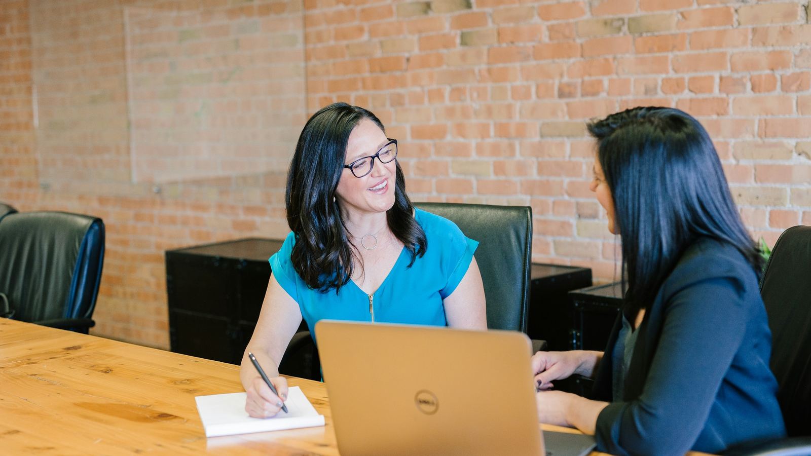 Two women talk across a desk with a laptop open